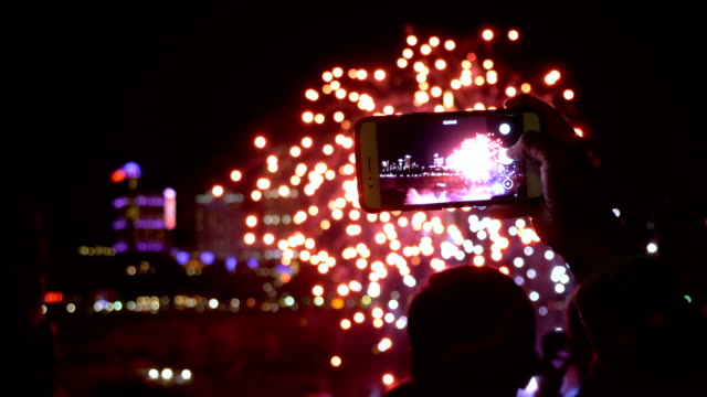 Human-hand-holding-smartphone-filming-a-video-of-night-fireworks,-people-and-buildings-in-the-background-slow-motion.