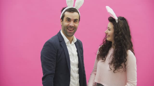 Young-lovers-couple-on-the-pink-background.-With-hackneyed-ears-on-the-head.-During-this,-the-girl-wears-rabbit-ears-to-her-husband.-After-that,-they-begin-to-make-a-photo-of-sephi-on-the-phone.-Very-smiling..