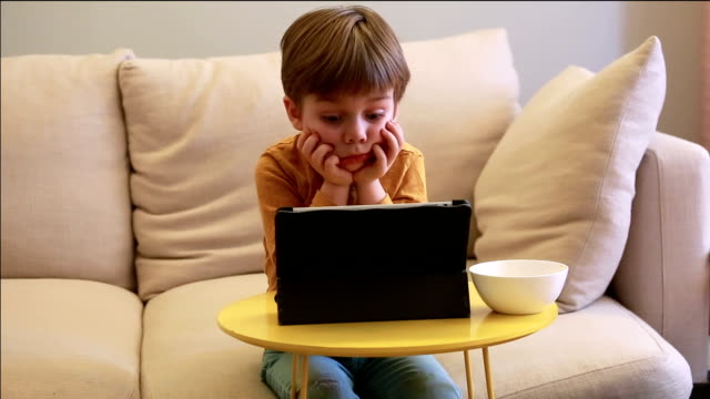 Kind-mit-Tablett-PC-auf-Bett-zu-Hause.-Niedlicher-Junge-auf-dem-Sofa-ist-zu-beobachten-Cartoon,-spielen-Spiele-und-lernen-von-Laptop.-Bildung,-Spaß,-Freizeit,-Glück,-moderne-Computertechnik-und-Kommunikation.
