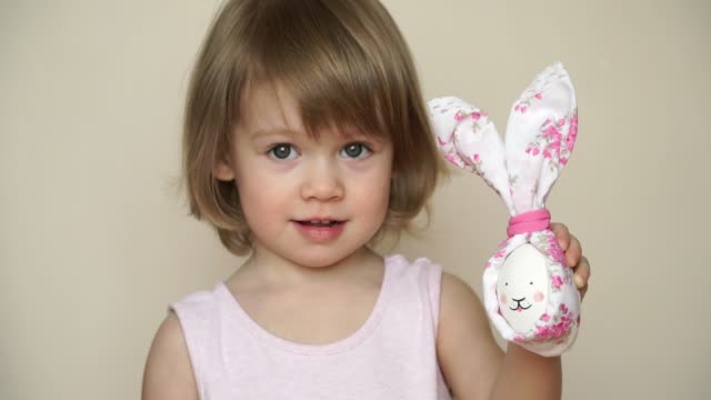 Portrait-of-little-cute-smiling-blonde-girl-holds-chicken-egg-decorated-for-Easter-bunny,-with-painted-muzzle-and-ears.