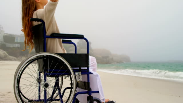 Seitenansicht-der-jungen-kaukasischen-Frau,-die-mit-Armen-sitzt,-die-auf-dem-Rollstuhl-am-Strand-4k-ausgestreckt-sind