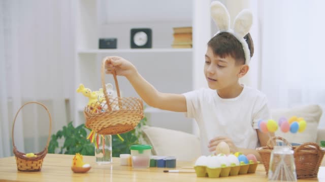 Cute-little-boy-with-bunny-ears-is-holding-a-wooden-basket,-full-of-Easter-colorful-eggs,-and-showing-with-his-finger-how-much-he-loves-Easter-conception.