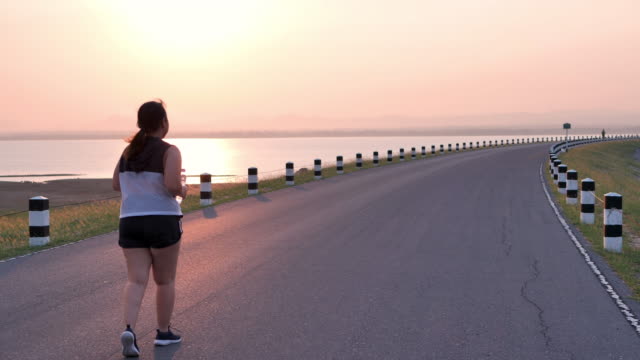 Übergewichtige-asiatische-Frauen-joggen-am-frühen-Morgen-im-Sonnenlicht-auf-der-Straße.-Konzept-des-Gewichtsverlustes-mit-Bewegung-für-die-Gesundheit.-Langsame-Bewegung,-Rückansicht