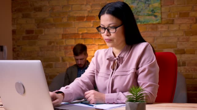 Closeup-shoot-of-adult-asian-businesswoman-working-on-the-laptop-and-handling-graphs-to-the-businessman-sitting-the-couch-and-using-the-tablet-on-the-background
