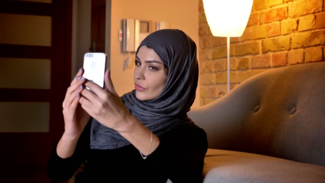 Nahauflage-Porträt-von-erwachsenen-attraktiven-muslimischen-Frauen-in-Hijab-nehmen-Selfies-am-Telefon,-während-auf-dem-Boden-in-der-Tür-in-einer-gemütlichen-Wohnung-sitzen
