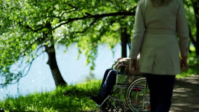 Tilt-down-of-disabled-senior-man-sitting-in-wheelchair-near-lake-in-park-alone,-looking-at-view-and-thinking.-Unrecognizable-woman-passing-by-on-windy-day