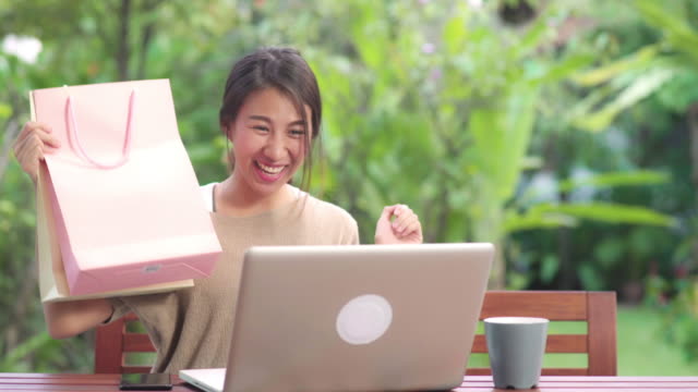 Asian-woman-using-laptop-video-conference-with-friends,-female-relax-feeling-happy-showing-shopping-bags-sitting-on-table-in-the-garden-in-morning.-Lifestyle-women-relax-at-home-concept.