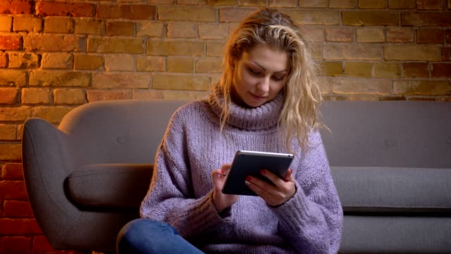 Closeup-top-view-shoot-of-adult-caucasian-blonde-female-using-the-tablet-looking-at-camera-and-laughing-while-sitting-on-the-floor-indoors-in-a-cozy-apartment