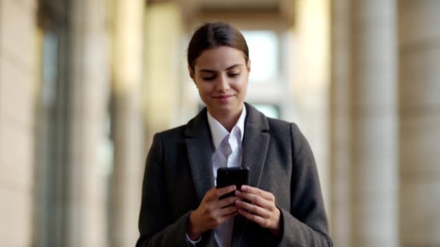 Waist-up-shot-of-attractive-young-woman-in-coat-walking-down-street-after-work.-Smiling-businesswoman-reading-text-messages-on-cell-phone-or-browsing-social-networks