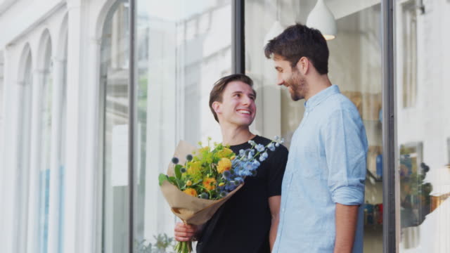 Pareja-gay-masculina-amorosa-sosteniendo-las-manos-saliendo-de-floristas-sosteniendo-un-montón-de-flores