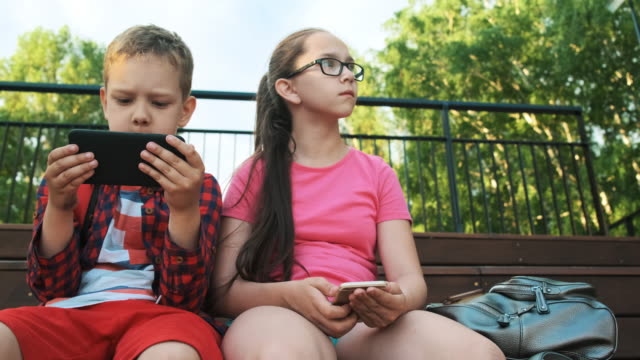 Die-ältere-Schwester-und-der-Bruder-sitzen-auf-der-Straße-und-schauen-auf-Smartphones.