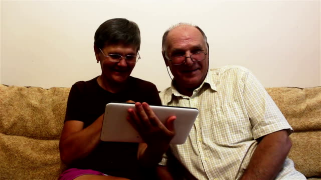 Ein-älteres-Ehepaar-sitzt-zu-Hause-auf-einem-Sofa-und-schaut-sich-Fotos-auf-einem-Tablet-PC-an,-lächelt-und-redet-aktiv.