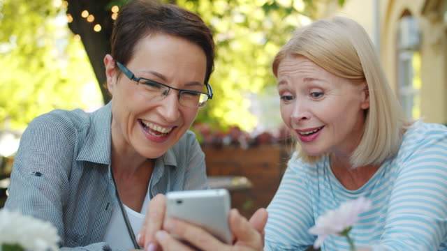Weibliche-Freunde-mit-Smartphone-in-Straßencafé-Blick-auf-Bildschirm-Spaß-haben