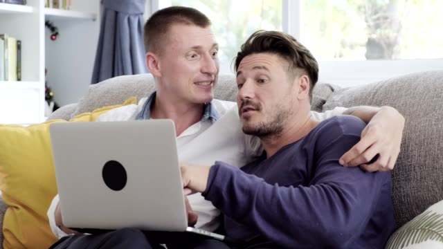 Gay-couple-relaxing-on-couch-using-laptop-computer.-Talking-exciting.