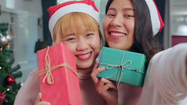Pareja-lesbiana-asiática-celebran-festival-de-Navidad.-LGBTQ-adolescente-mujer-relajarse-feliz-sosteniendo-regalo-y-el-uso-de-selfie-smartphone-con-árbol-de-Navidad-disfrutar-de-las-vacaciones-de-invierno-de-Navidad-en-la-sala-de-estar-en-casa.