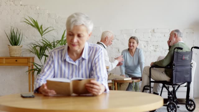 Rack-focus-of-three-elderly-people,-two-men-including-disabled-one-on-wheelchair-and-woman,-playing-cards-and-talking-in-nursing-home.-Intelligent-old-woman-reading-book-sitting-at-table-on-foreground