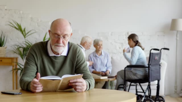 Rack-Fokus-von-drei-älteren-Menschen,-zwei-Frauen-einschließlich-behinderter-und-Mann,-Karten-im-Pflegeheim-spielen.-Senior-Mann-in-Brille-lesen-Buch,-Blick-in-die-Kamera-und-lächelnd-sitzend-am-Tisch