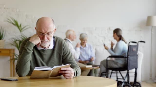 Rack-Fokus-von-drei-älteren-Menschen,-zwei-Frauen-einschließlich-behinderter-und-Mann,-Karten-im-Pflegeheim-spielen.-Intelligenter-Senior-in-Brille-lesen-Buch-und-denken-sitzen-am-Tisch