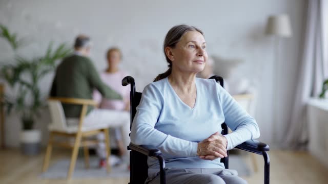 Tracking-shot-of-disabled-senior-woman-in-wheelchair-looking-away-thoughtfully,-then-turning-head-and-smiling-at-camera-happily-in-nursing-home,-other-aged-patients-in-background