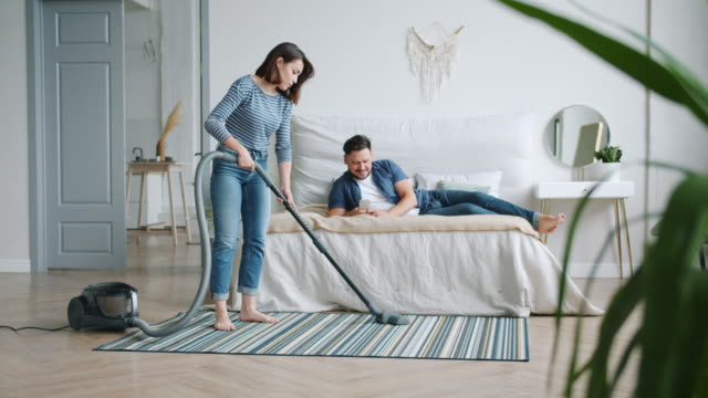 Housewife-cleaning-floor-with-vacuum-cleaner-while-man-using-smartphone-in-bed