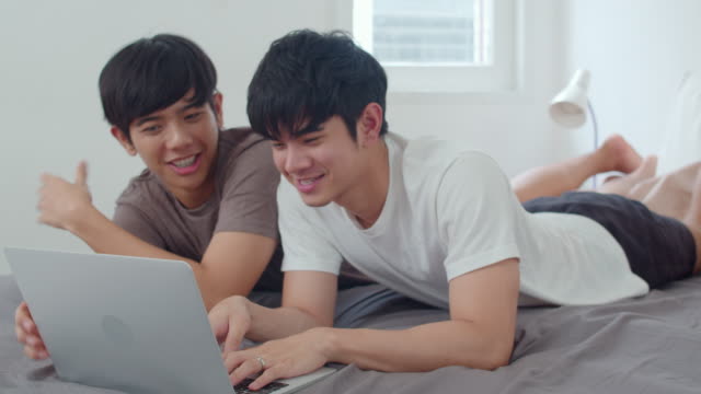 Asian-Gay-lgbtq-men-couple-using-computer-laptop-at-modern-home.-Young-Asia-lover-male-happy-relax-rest-together-after-wake-up,-watching-movie-lying-on-bed-in-bedroom-at-house-in-the-morning-concept.
