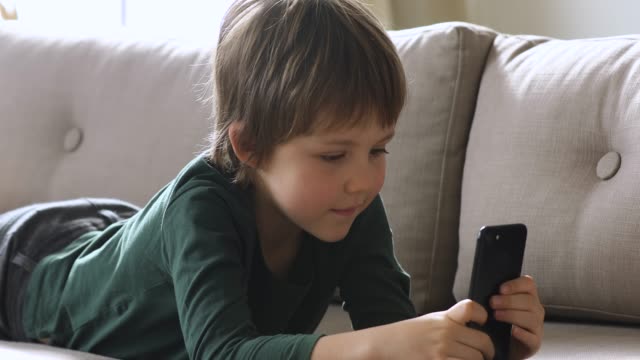 Curious-cute-schoolboy-holding-smartphone-watching-videos-alone-at-home