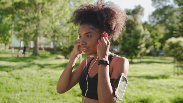 Glückliche-Athletin-afrikanische-amerikanische-Läuferin-setzen-Kopfhörer-in-ihre-Ohren-hören-Musik-auf-Smartphone-in-Armbinde-im-Park