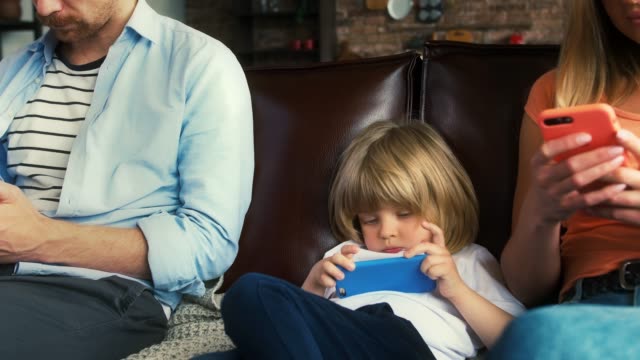 Der-junge-Vater,-die-Mutter-und-ihr-Kleiner-in-Freizeitkleidung-nutzen-Smartphones,-während-sie-im-Wohnzimmer-auf-der-Couch-sitzen.-Nahaufnahme,-Zeitlupe