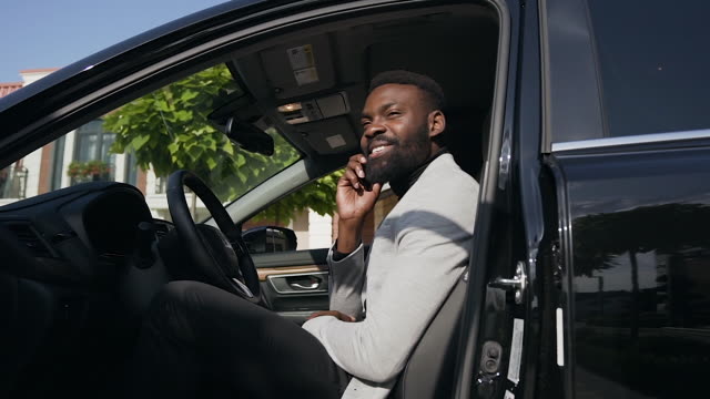 Young-African-businessman-in-suit-sitting-at-wheel-of-new-car-at-parking-near-home-while-talking-on-smartphone.-Dolly-shot