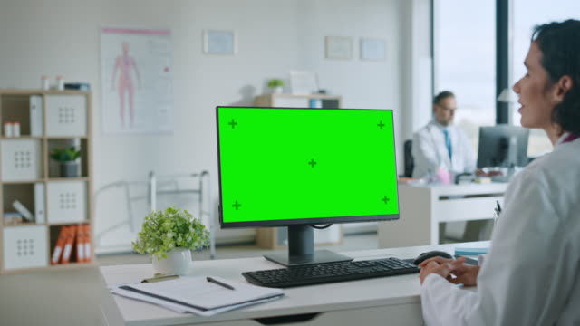 La-doctora-médica-está-haciendo-una-videollamada-con-el-paciente-en-un-ordenador-con-pantalla-de-simulación-de-pantalla-verde-en-una-clínica-de-salud.-Assistant-in-Lab-Coat-está-hablando-sobre-problemas-de-salud-en-la-oficina-del-hospital.