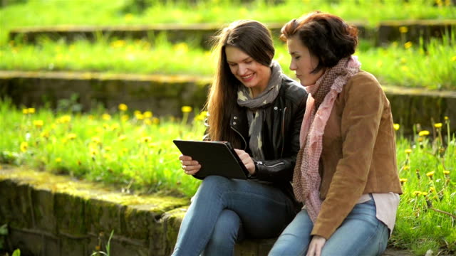 Young-Women-with-Toothy-Smiles-are-Using-Tablet-in-Hands.-Attractive-Brunettes-are-Having-Fun-Together-with-Electronic-Device-in-the-Park-during-Sunny-Spring-Day-Outdoors