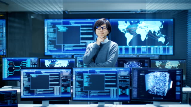 In-the-System-Control-Room-Smart-Engineer-Turns-and-Makes-Thinking-Gesture.-High-Tech-Facility-Has-Multiple-Monitors-with-Graphics.