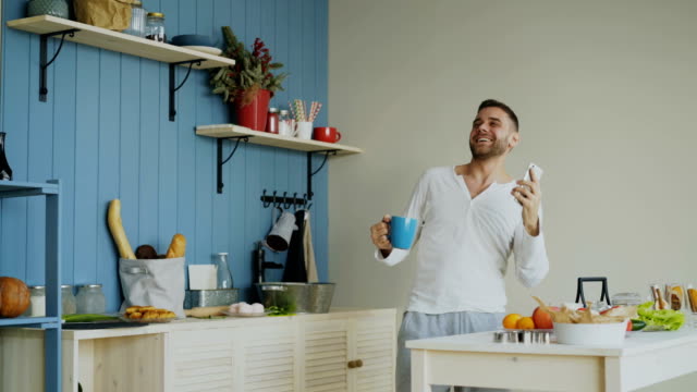 Cheerful-happy-man-dancing-and-singing-in-kitchen-while-using-his-smartphone-at-home-in-the-morning