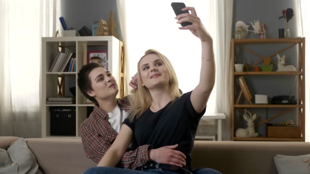 Zwei-junge,-schöne-Mädchen-sitzen-auf-der-Couch-und-machen-Selfies,-Lesben,-LGBT,-blond,-brünett.-60fps