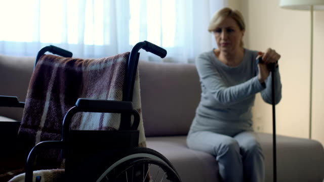 Depressed-female-pensioner-sitting-on-couch-and-looking-at-wheelchair,-rehab