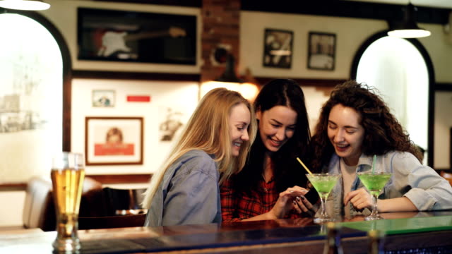 Girls-are-using-smartphone,-laughing-and-talking-while-sitting-in-bar-together.-Girls-in-casual-clothing-are-watching-screen-and-chatting.-Modern-technologies-for-fun-concept.