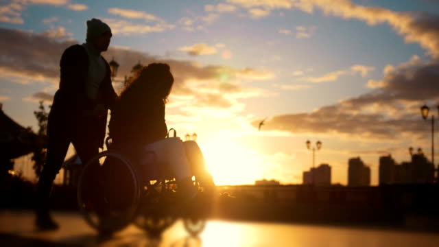 Fürsorglichen-Mann-mit-einer-behinderten-Frau-im-Rollstuhl-zu-Fuß-durch-Kai-bei-Sonnenuntergang
