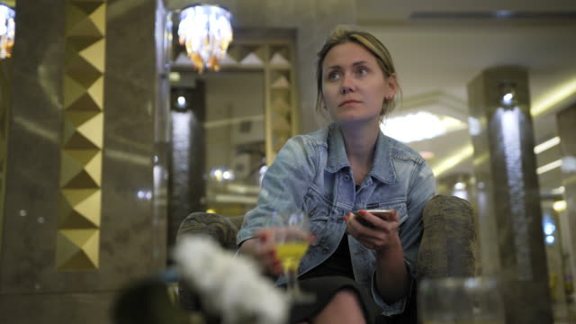 young-Caucasian-woman-in-a-jeans-jacket-drinks-juice-from-a-glass-and-uses-a-smartphone,-writes-messages-on-the-social-network.-Sits-at-the-table-in-the-hotel-restaurant.-Concept-of-healthy-fresh-food-in-business.-transfer-focus-from-foreground-to