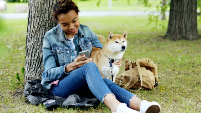 Happy-African-American-woman-is-using-smartphone-and-caressing-her-cute-pet-dog-resting-in-city-park-on-windy-summer-day.-Nature,-animals-and-people-concept.