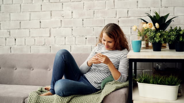 Attraktives-Mädchen-in-Freizeitkleidung-nutzt-Smartphone-auf-Sofa-im-modernen-Loft-Stil-Wohnung-sitzen-und-Lächeln-auf-den-Lippen.-Moderne-Technologie,-innen--und-Jugend-Konzept.