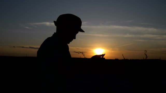 Silhouette-eines-jungen-verwendet-eine-Tablette-bei-Sonnenuntergang-im-Feld-liest-etwas-auf-dem-Tablet-im-freien