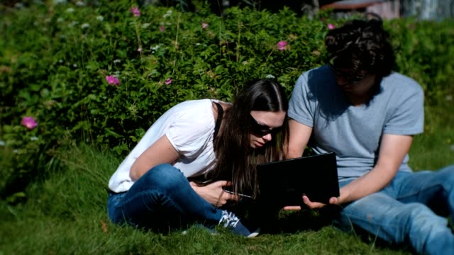 Young-couple-man-and-woman-are-working-together-on-mobile-and-laptop-sitting-in-park.