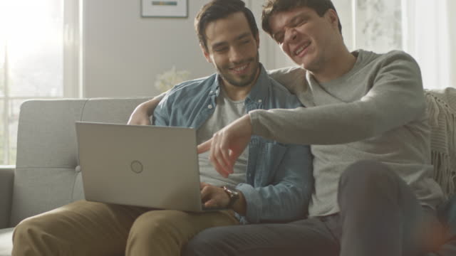 Cute-Male-Gay-Couple-Spend-Time-at-Home.-They-Sit-on-a-Sofa-and-Use-the-Laptop.-They-Browse-Online.-Partner's-Hand-is-Around-His-Lover.-They-Give-Each-Other-a-High-Five.-Room-Has-Modern-Interior.