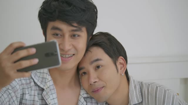 Junge-asiatische-schwules-Paar-sitzt-am-Bett-Umarmung-und-Telefon-unter-Selfie-zusammen-Schlafzimmer-zu-Hause-benutzen.-Lebensstil-LGBT-paar-gemeinsam-im-Haus-Konzept.