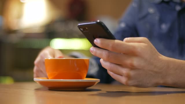 Nahaufnahme-der-Hände-eines-Mannes-mit-Smartphone-und-trinken-aus-einer-dampfenden-Tasse-Kaffee-in-einem-Café-in-Medellin-Kolumbien