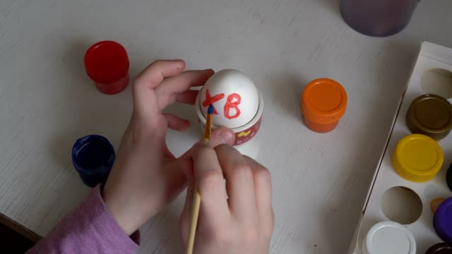 El-niño-pinta-los-colores-del-huevo-de-Pascua-y-dibuja-símbolos-en-él-con-un-cepillo.-De-cerca.-La-vista-desde-la-cima.-4k.