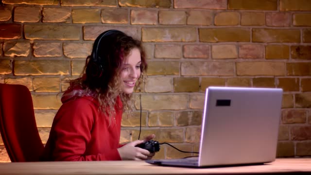 Porträt-der-jungen-Bloggerin-in-roten-Hoodie-spielen-Videospiel-mit-Joystick-und-gewinnen-glücklich-auf-bricken-Wand-Hintergrund.