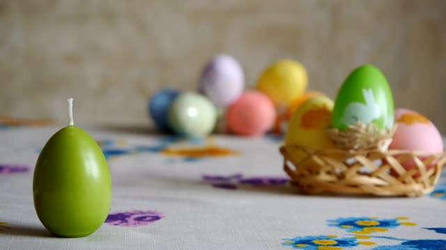 Mano-femenina-poniendo-en-la-mesa-tres-velas-de-Pascua.-Verde,-naranja-y-amarillo.-Coloridos-huevos-de-Pascua-en-el-fondo.