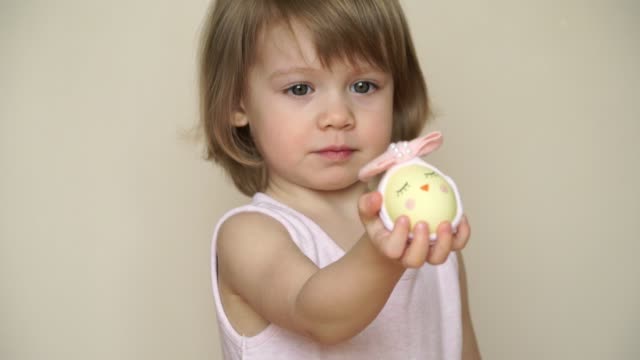 Portrait-of-little-cute-smiling-blonde-girl-holds-chicken-egg-decorated-for-Easter-chick,-painted-muzzle-and-bow.