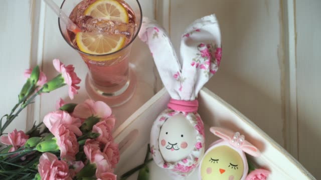 Close-up-Easter-eggs-decorated-for-Easter-bunny-and-Easter-chick-with-painted-muzzle,-against-background-of-pink-carnations-and-lemonade-with-lemon